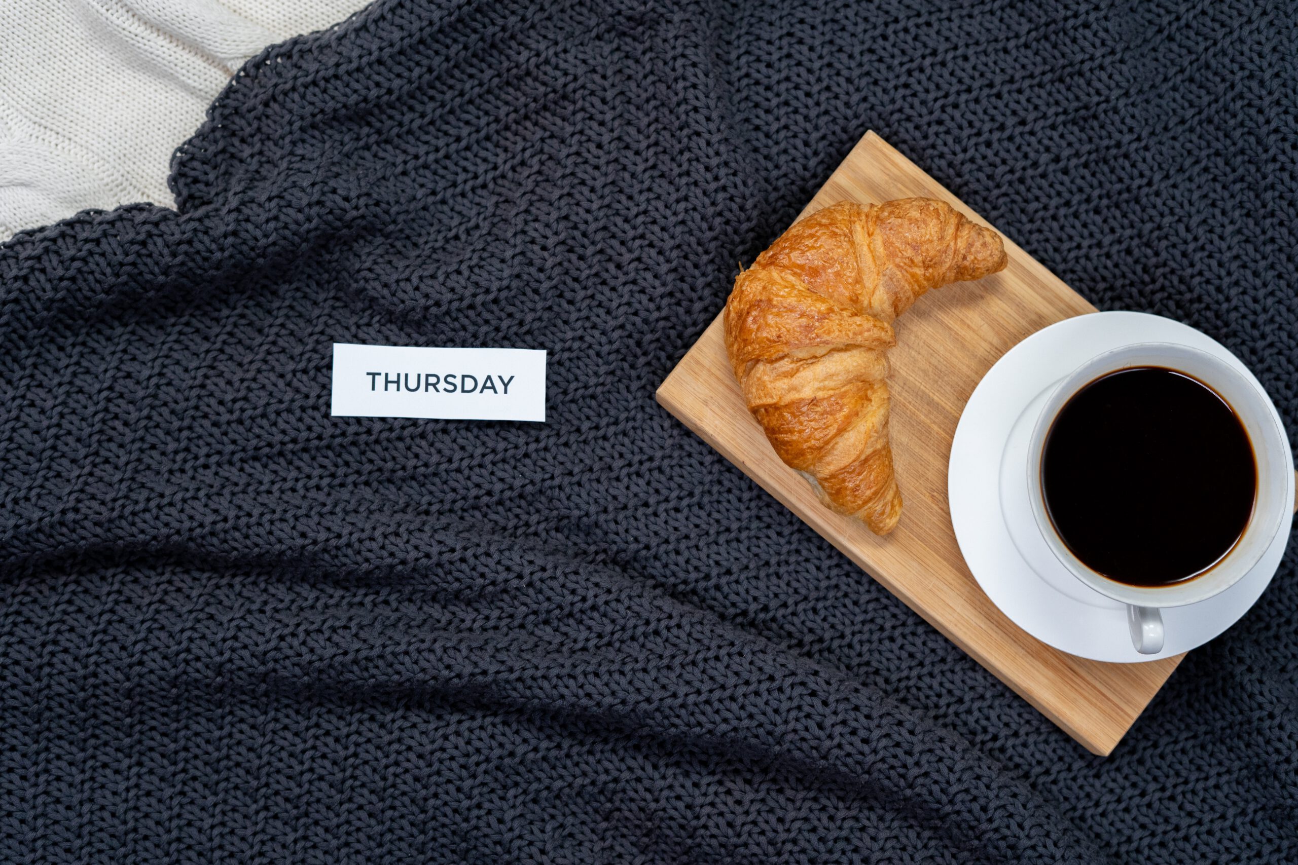 Karteczka z napisem czwartek leżąca na kocu obok tacy z kawą i croissantem